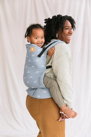 A smiling boy sitting in back-carry position in the Tula Harbor Skies Toddler Carrier.
