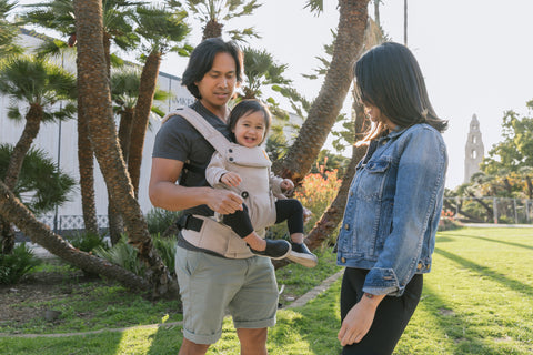 A father carrying his child in a forward-facing front carry position while being in warm weather.