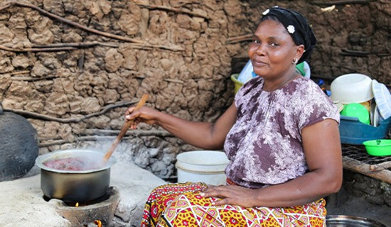 Surrounded by pots and bowls, a woman sits and uses a wooden spoon to stir food cooking in a pot. 