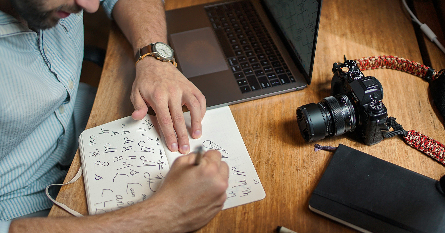 Ein Mann skizziert in ein Notizbuch verschiedene Logos. Vor ihm liegt eine Kamera und ein  Laptop.