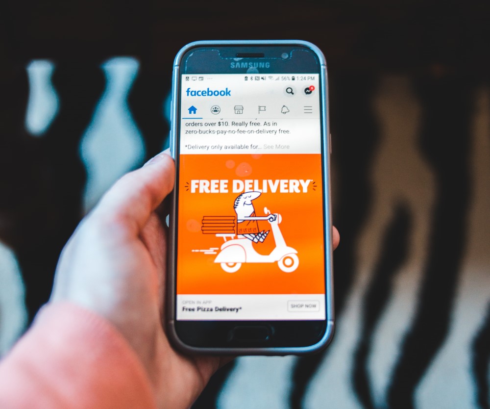 Eine Hand hält ein Handy, auf dem die Facebook Startseite mit einem Post zu kostenloser Pizza Lieferung geöffnet ist.