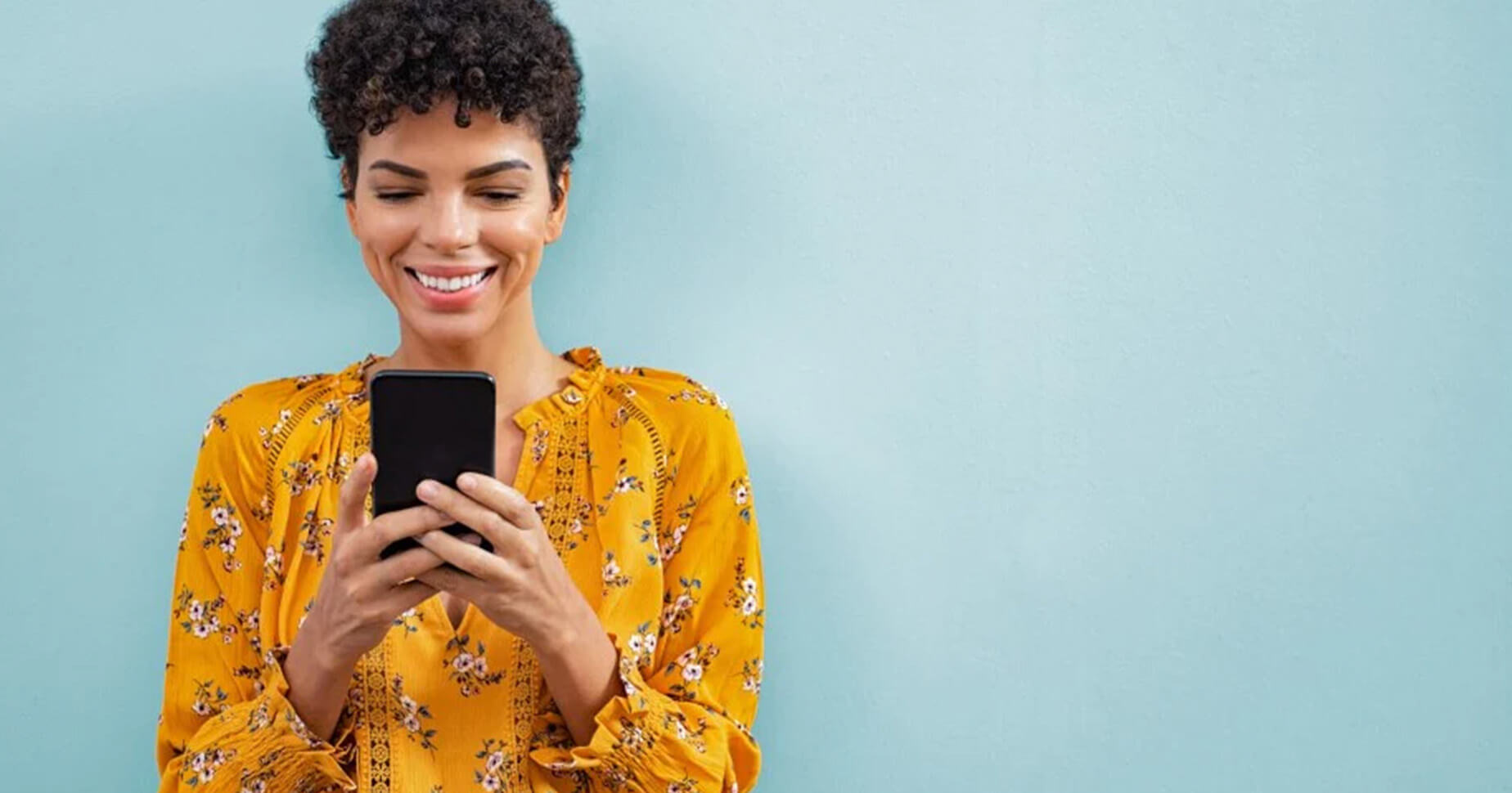 Eine Frau steht vor einem hellblauen Hintergrund und schaut lächelnd auf ihr Handy. Im Blogbeitrag erfährst du, wie du Facebook Pixel richtig nutzt.
