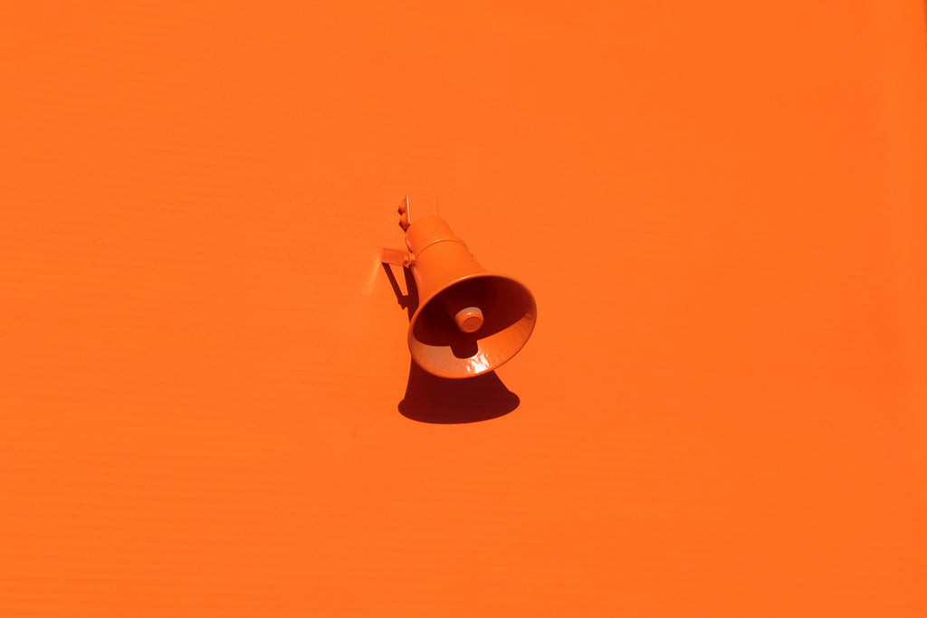 Das Foto zeigt einen orangen Hintergrund auf dem ein Megaphon in derselben Farbe zu sehen ist.