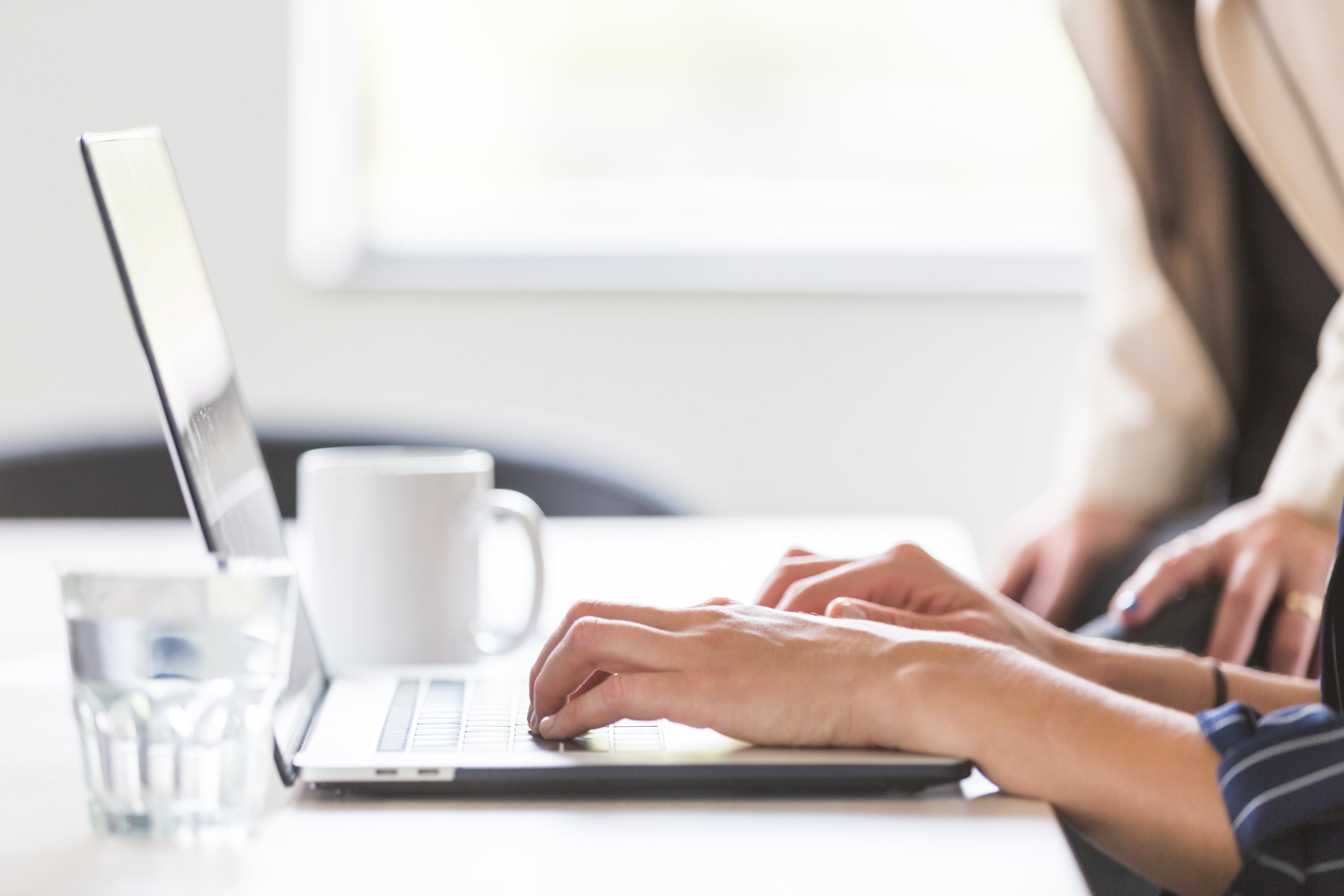 Blick von der Seite auf einen Schreibtisch auf dem ein Laptop neben einem Glas Wasser und einer Kaffeetasse steht. Eine Person tippt gerade etwas, während eine zweite Person über die Schulter guckt. Wir zeigen dir, welche typischen Fehler du beim Erstellen eines Businessplans vermeiden solltest!