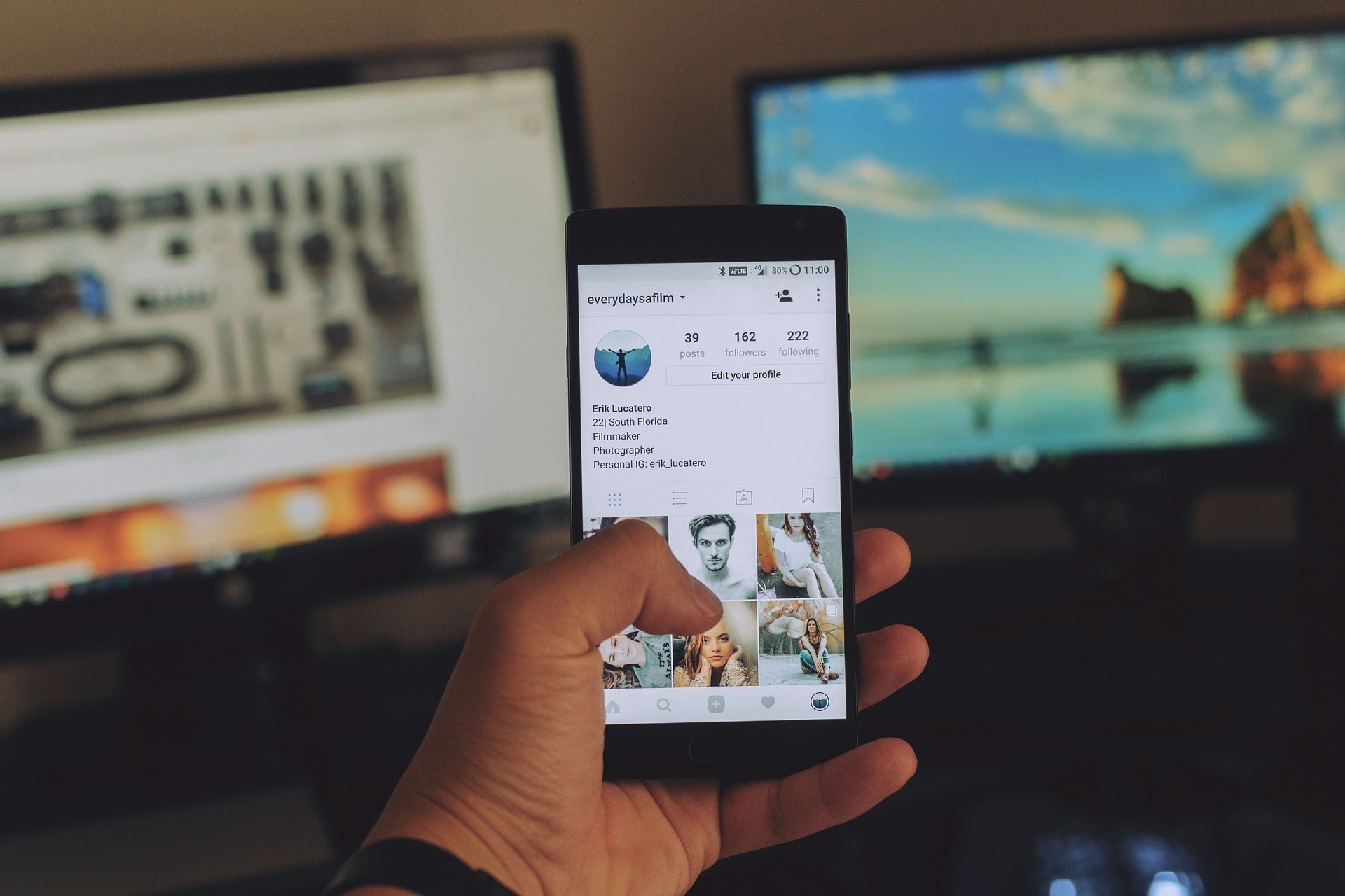 Blick auf ein Handy in der Hand einer Person. Auf dem Bildschirm ist ein Instagram-Profil zu sehen. Im Hintergrund stehen zwei Bildschirme. Danke an Kund:innen kannst du auch sagen, indem du nutzergenerierte Beiträge auf deinen Social-Media-Profilen postest.