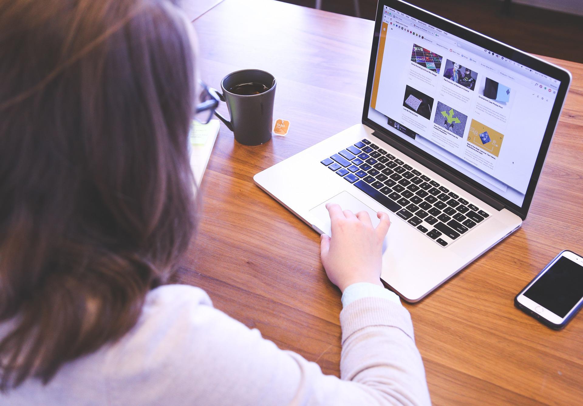 Blick auf einen aufgeklappten Laptop auf einem Tisch neben einer Kaffeetasse und einem Handy. Eine Frau sitzt mit dem Rücken zum Betrachter gewandt vor dem Laptop und scrollt mit der rechten Hand auf dem Trackpad.