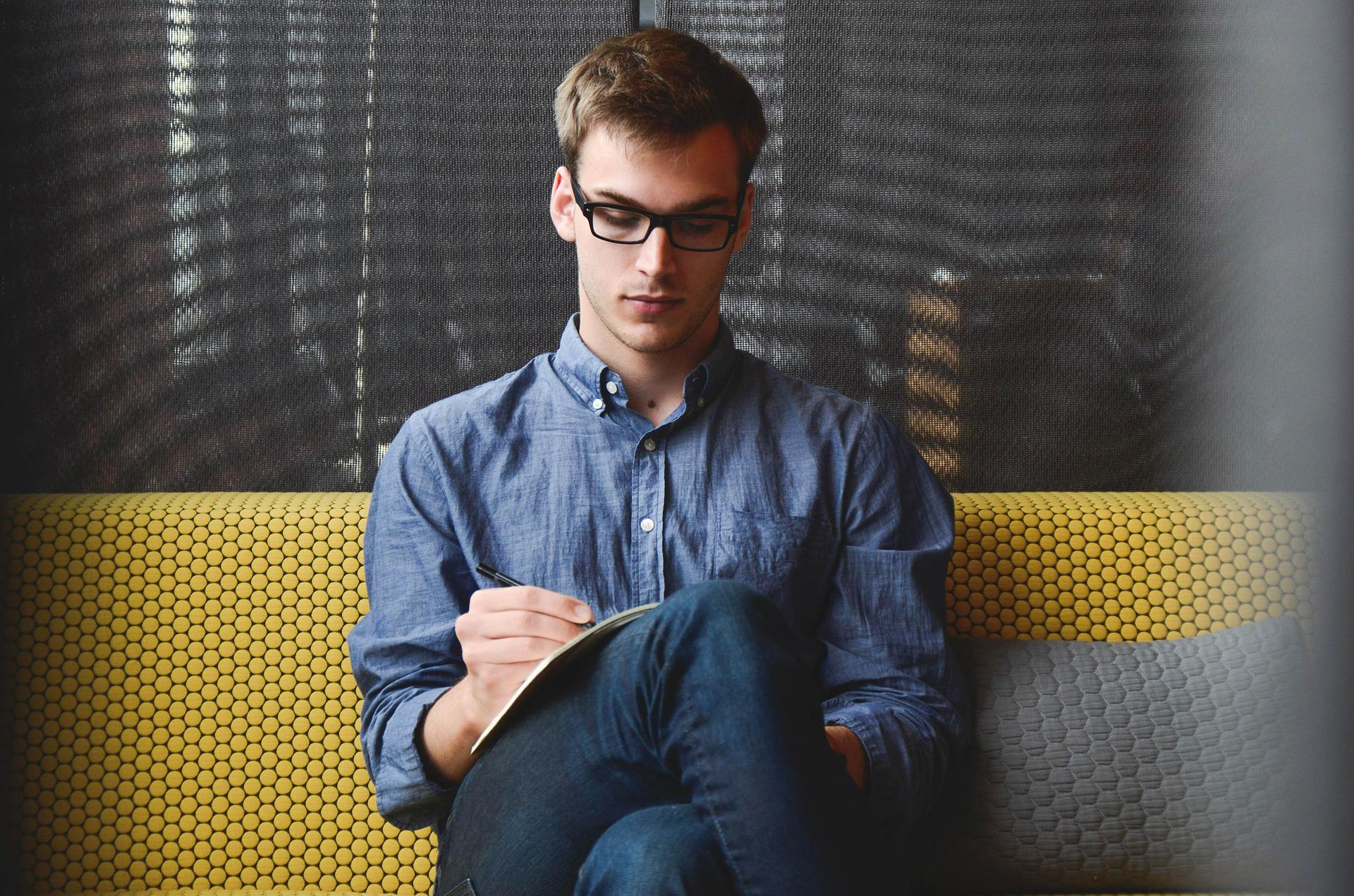 Ein junger Mann mit Brille in einem blauen Hemd sitzt auf einem gelbem Sofa mit einem Notizbuch auf dem Schoß und einem Stift in der rechten Hand.