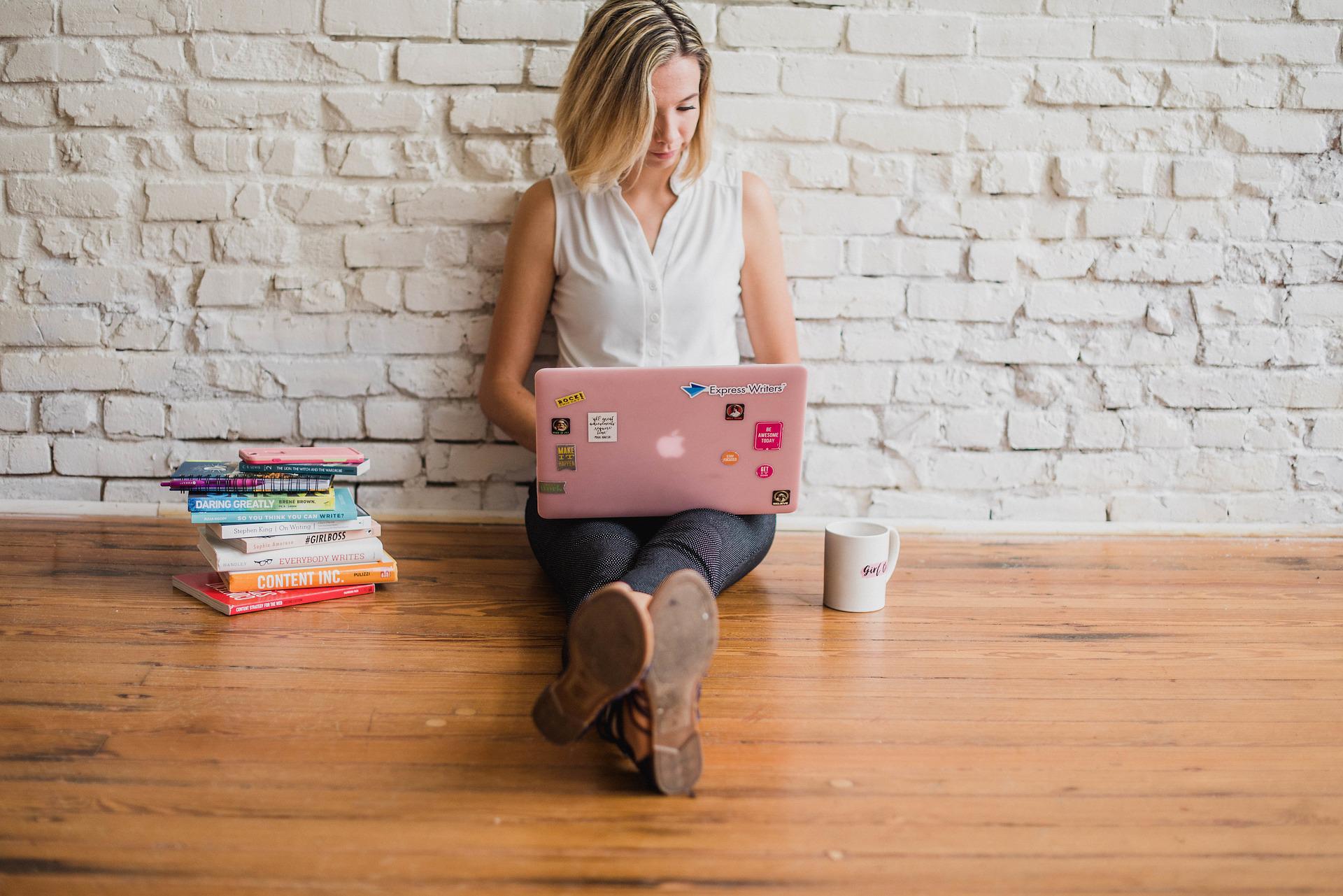 Eine junge Frau mit aufgeklappten Laptop auf dem Schoß sitzt vor einer gekachelten Wand auf dem Boden. Neben ihr steht links ein Stapel Bücher und rechts eine Kaffeetasse.