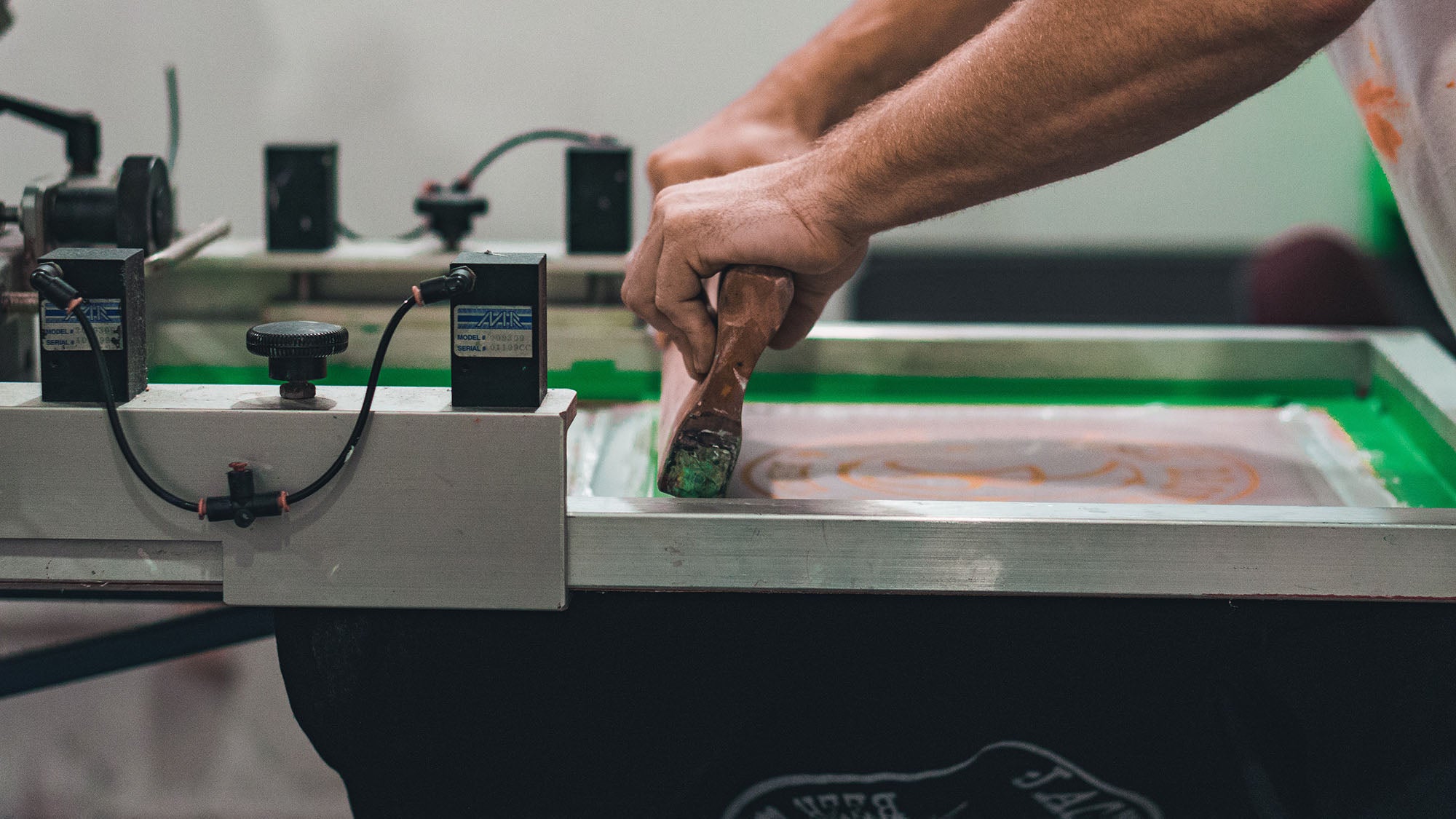 Ansicht eines Siebdruck-Geräts von der Seite an der eine Person arbeitet. T-Shirts und andere Druckartikel sind eine gute Grundlage für dein DIY-Geschäft. Egal ob Print-on-Demand oder eigene Herstellung über Siebdruck – es gibt unzählige Möglichkeiten. Mehr Tipps für dein DIY-Business findest du in diesem Beitrag!