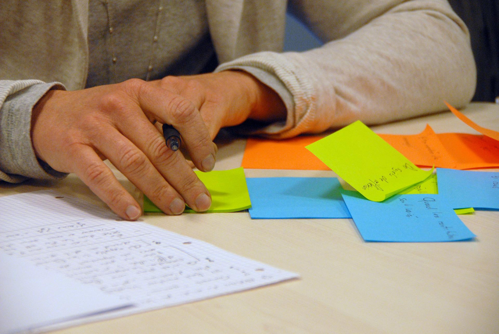 Blick auf einen Schreibtisch an dem eine Person sitzt. Es sind nur die Hände zu erkennen, die vor einem Stapel Notizzettel und Papier liegen.