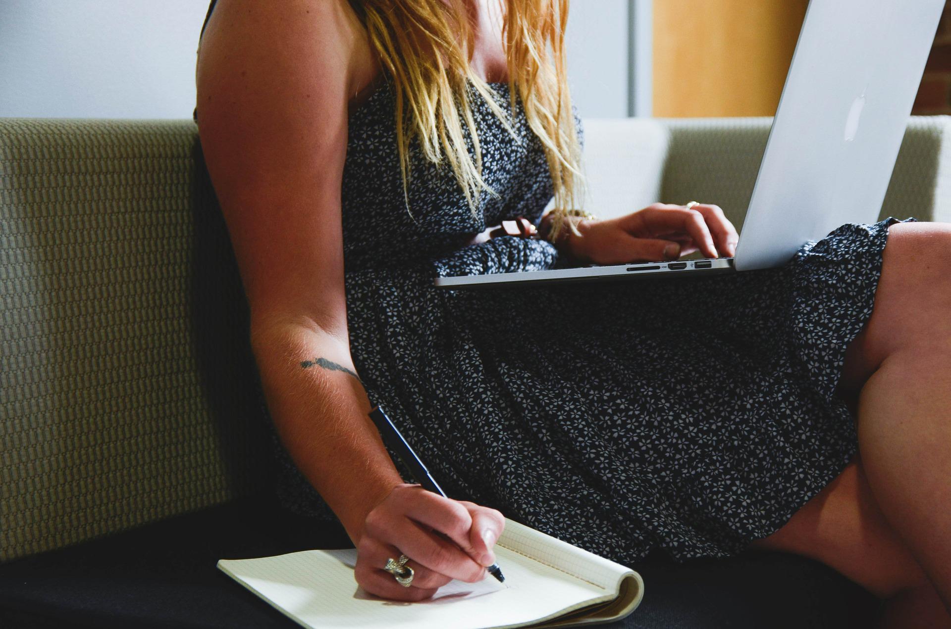 Mit Dropshipping Geld verdienen: Eine Frau sitzt mit Laptop auf dem Schoß auf einem Sofa. Rechts neben ihr liegt ein Notizbuch, in das sie etwas hineinschreibt.