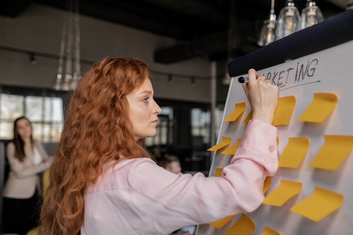 Eine Frau steht vor einem Whiteboard auf dem gelbe Post-Its angebracht sind.