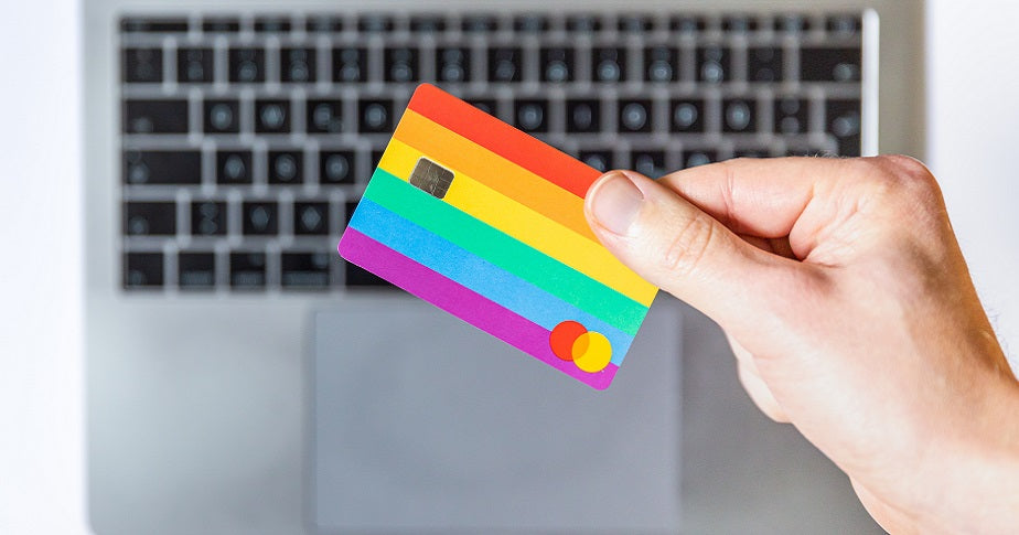 Eine Person hält eine regenbogenfarbene Kreditkarte vor einem Laptop ins Bild. 