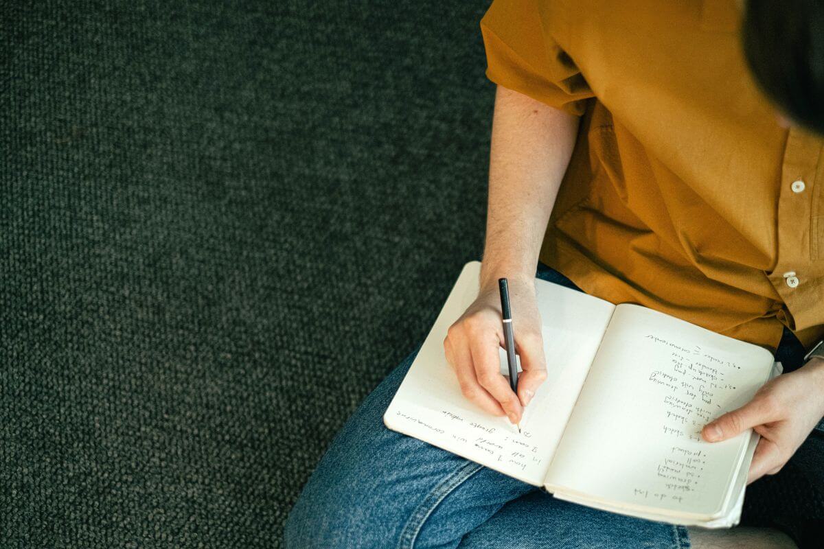 EIn Mann mit gelbem Hemd, der auf einem Sofa sitzt und in sein Notizbuch schreibt, wie man Schritt-für-Schritt eine GmbH gründet