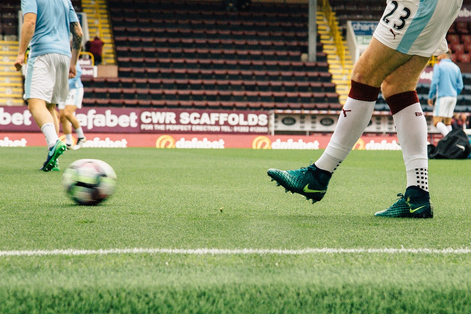 Zwei Sportler:innen im Trikot spielen sich auf einem Fußballplatz den Ball zu.