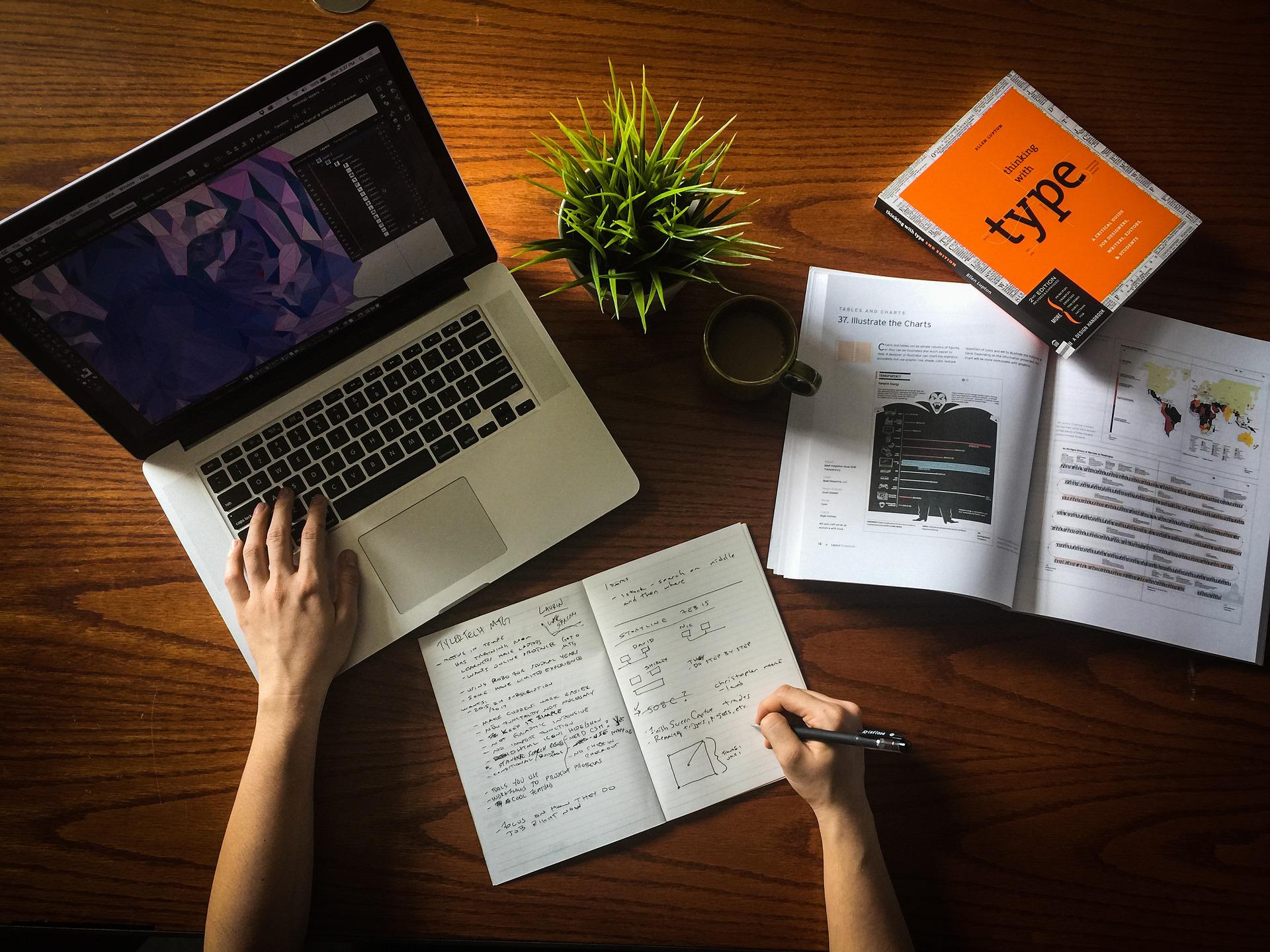 Blick von oben auf einen Schreibtisch auf dem ein aufgeklappter Laptop, eine Zimmerpflanze, zwei Bücher und ein Notizbuch liegen. Eine Person bedient mit der linken Hand den Laptop und schreibt mit der rechten Hand in das Notizbuch. Die Auswahl der richtigen Geschäftsidee ist entscheidend, um erfolgreich Haustierprodukte zu verkaufen.