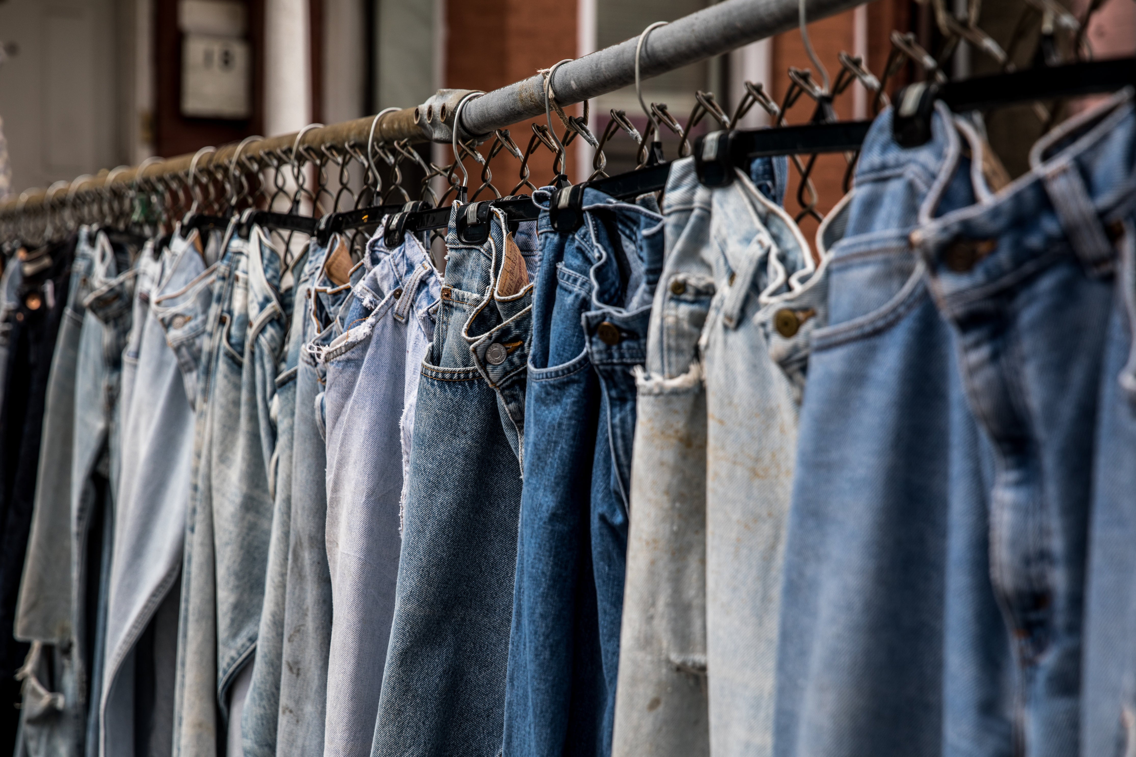 Eine Reihe von Jeanshosen in unterschiedlichen Farben hängt auf einer verrosteten Stange. Kleidung produzieren lassen bringt einige Herausforderungen mit sich. Wir zeigen dir, welche das sind!