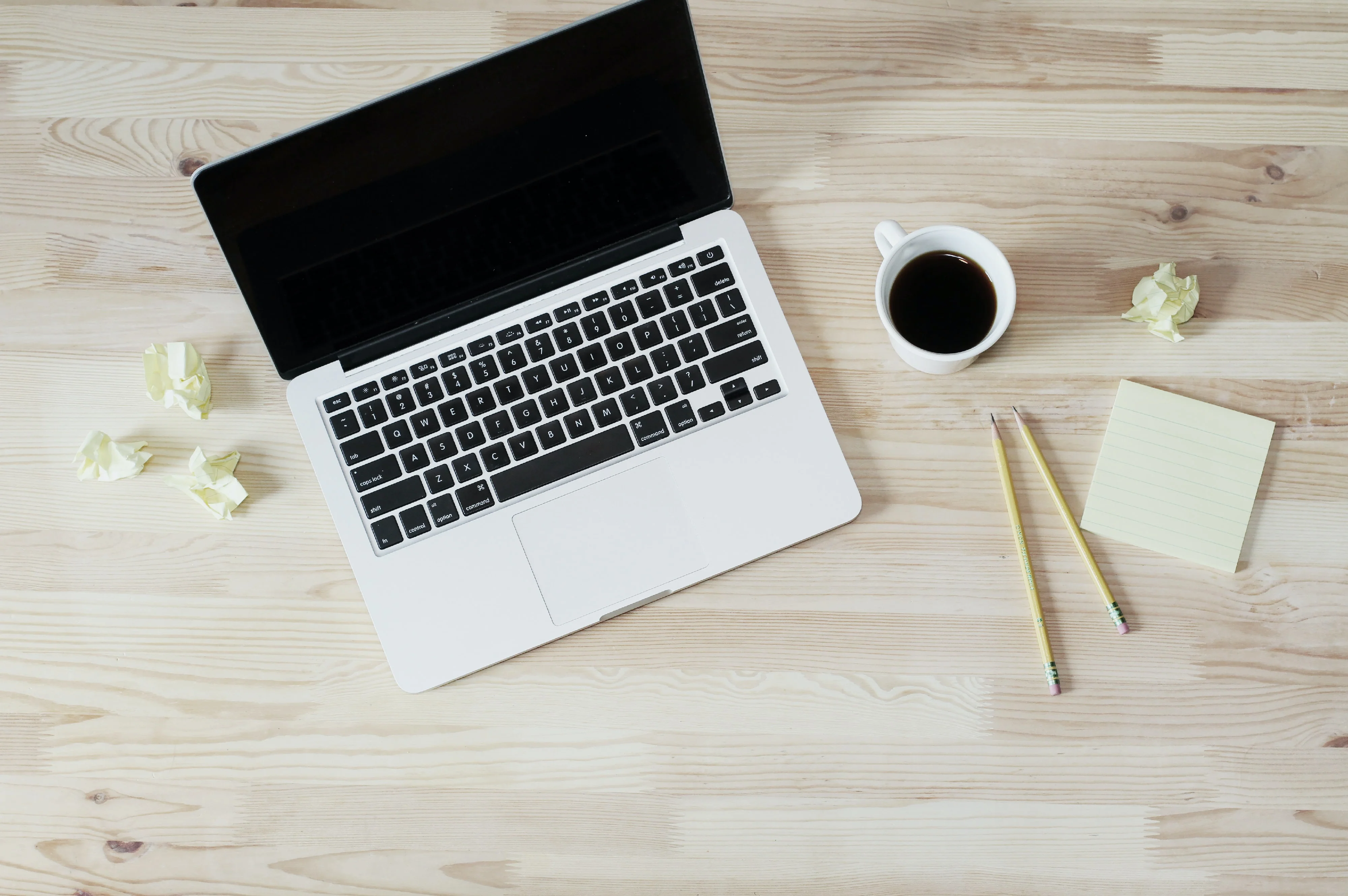 Blick von oben auf einen Schreibtisch auf dem ein aufgeklappter Laptop neben einer Tasse Kaffee sowie einem Notizblock und zwei Stiften liegt. Auf dem Tisch liegen verstreut zusammengeknüllte Notizen. Bevor du Kleidung produzieren lassen und ein Modelabel gründen kannst, musst du einige Vorbereitungen treffen.