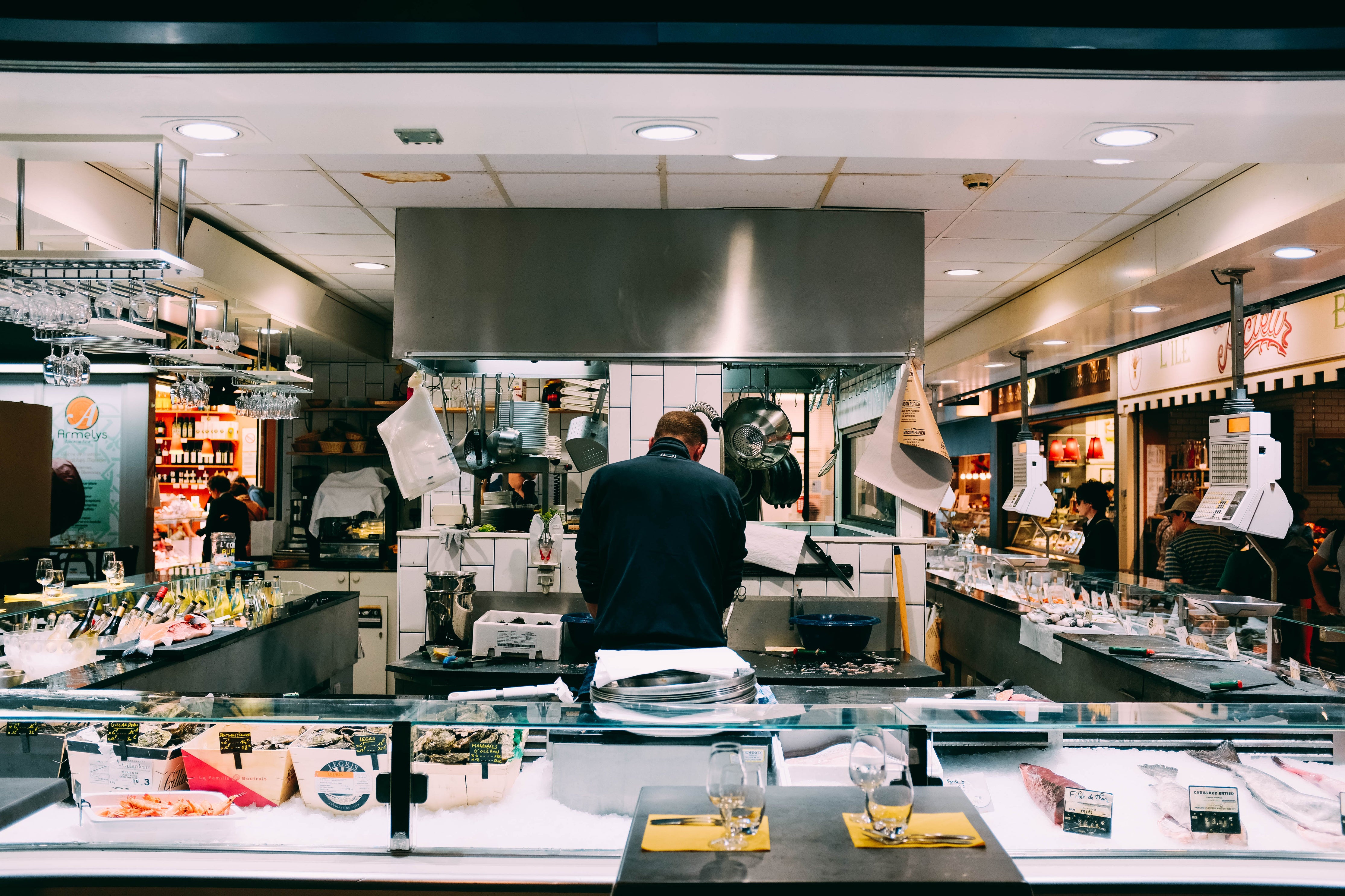 Mit dem Rücken zur Kamera ist dieser Koch mit der Zubereitung von Speisen in einem Restaurant mit offener Küche beschäftigt. Die gläsernen Arbeitsflächen im Zentrum eines Einkaufszentrums ermöglichen den Passanten einen Blick auf die köstlichen Speisen und Weine. Wenn du Lebensmittel verkaufen möchtest, musst du strenge Hygienevorschriften beachten.