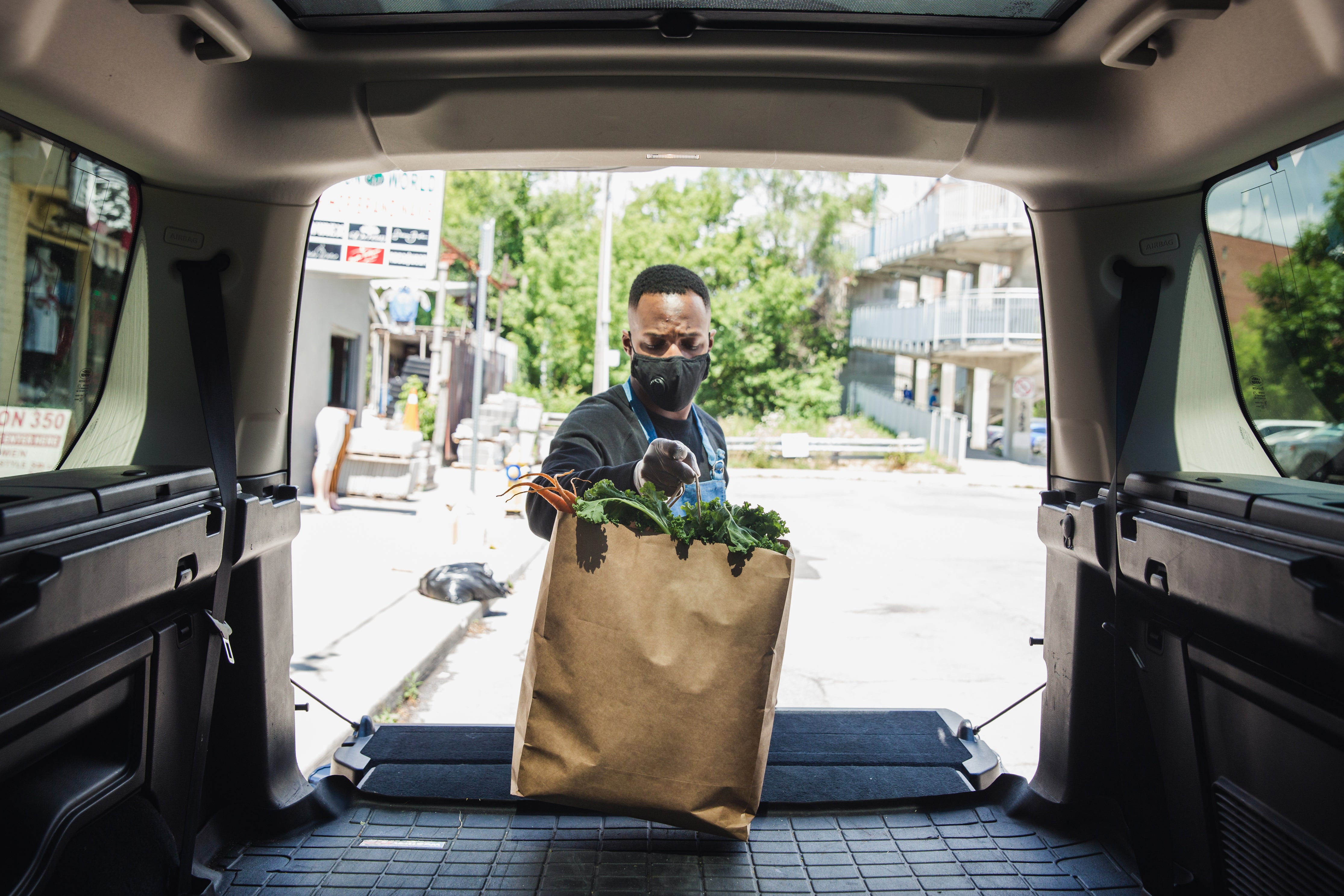 Blick aus dem Kofferraum eines Transporter auf einen Mann mit Maske, der gerade eine Tüte voller Lebensmittel in den Transporter stellt. Lebensmittel verkaufen geht bequem online. Falls der Versand nicht möglich ist, kannst du alternative Optionen wie die Abholung vor Ort anbieten.