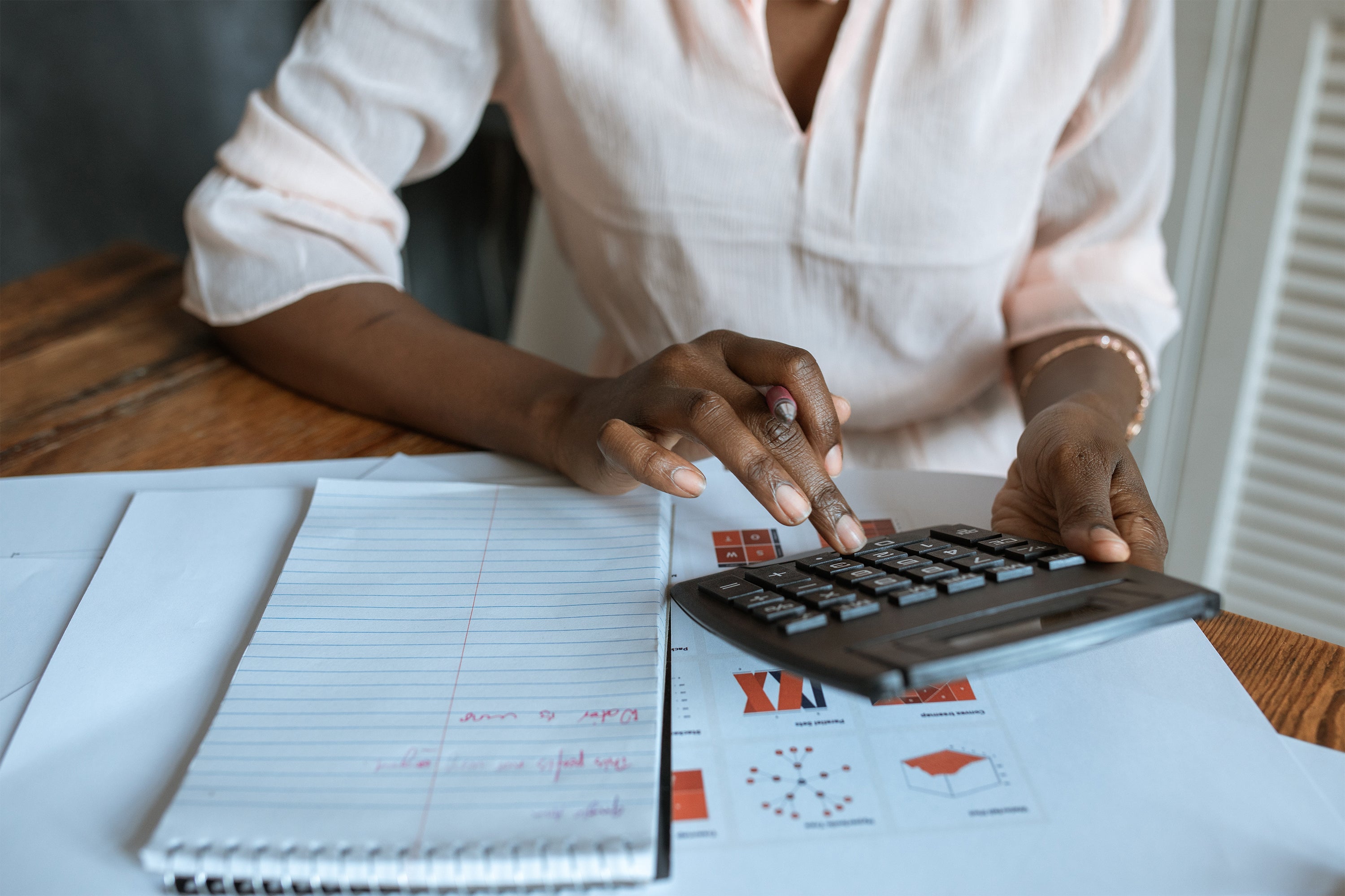 Eine Frau, die an einem Holztisch sitzt und mit einem Taschenrechner rechnet welche Steuern beim Nebengewerbe anmelden auf sie zukommen