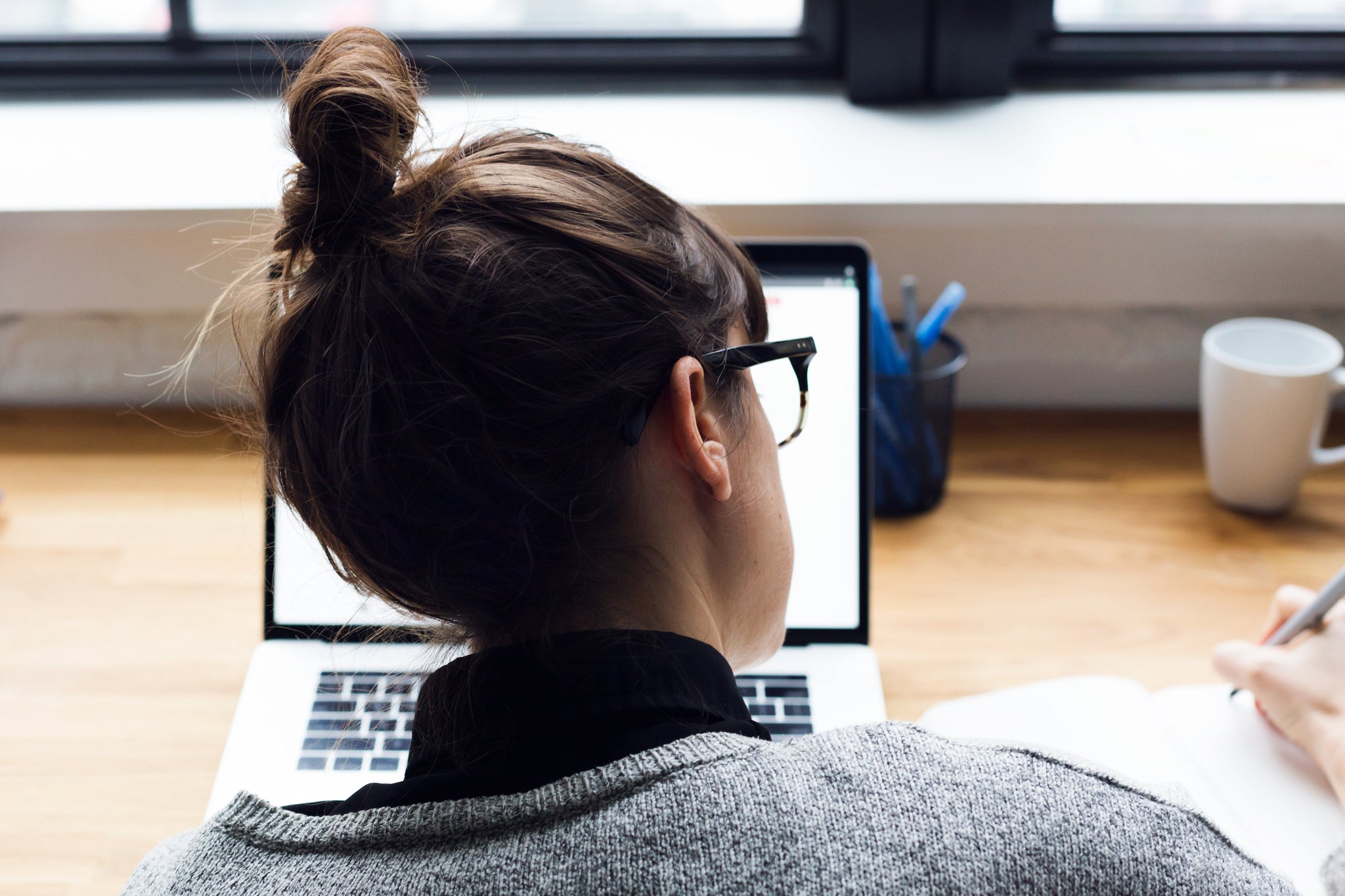 Eine junge Frau mit Brille sitzt an einem Tisch vor einem großen Fenster. Vor ihr steht ein Laptop. Während sie etwas darauf liest, macht sie sich Notizen in einem Notizbuch neben ihr. Einen Newsletter zu erstellen kann viele Vorteile haben. Wir zeigen dir welche!