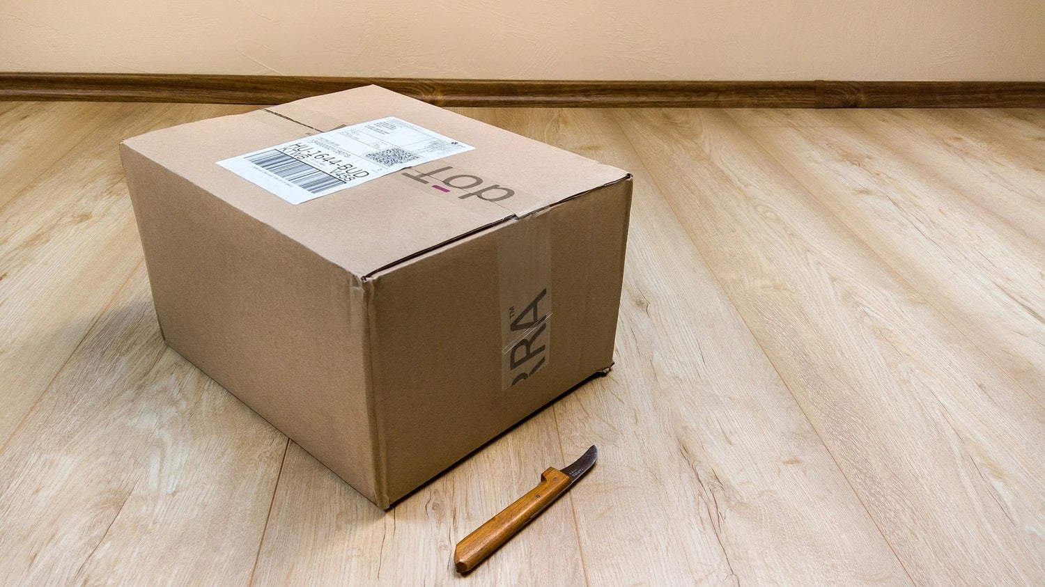 Blick auf ein gepacktes Paket, das neben einem Cutter auf einem Holzboden steht.