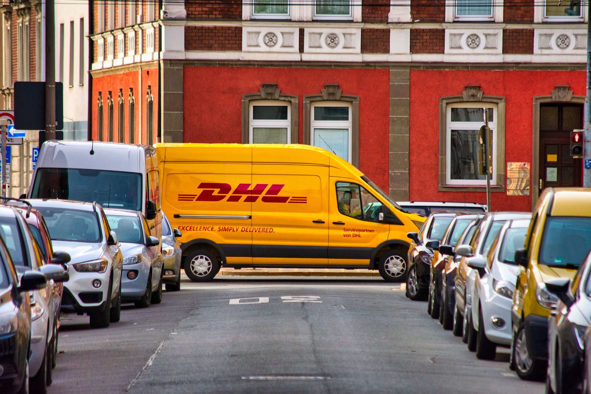 Blick in eine Straße auf der links und rechts Autos parken und am Ende ein DHL-Paketauto steht. Wir erklären dir Schritt für Schritt den Versandprozess und auf was du achten solltest, wenn du einen optimalen Onlineshop-Versand aufbauen willst! 