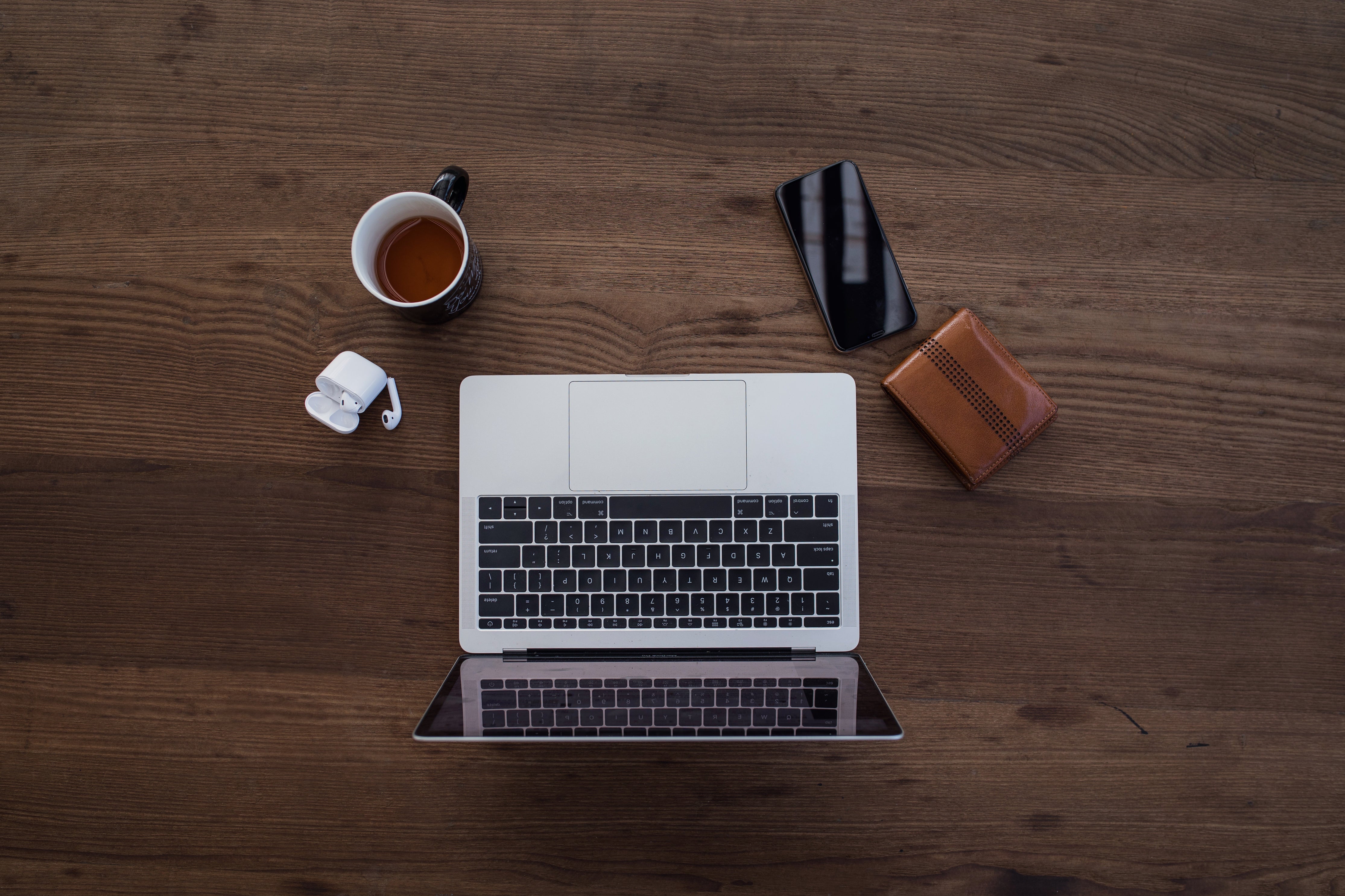 Blick von oben auf einen dunkelbraunen Tisch auf dem ein aufgeklappter Laptop steht. Neben dem Laptop steht links eine Tasse mit Kaffee und AirPods. Rechts daneben liegen ein Handy und eine Brieftasche. Der Online-Shop-Warenkorb ist der Einstieg in den Conversion-Funnel und sollte daher optimal gestaltet sein!