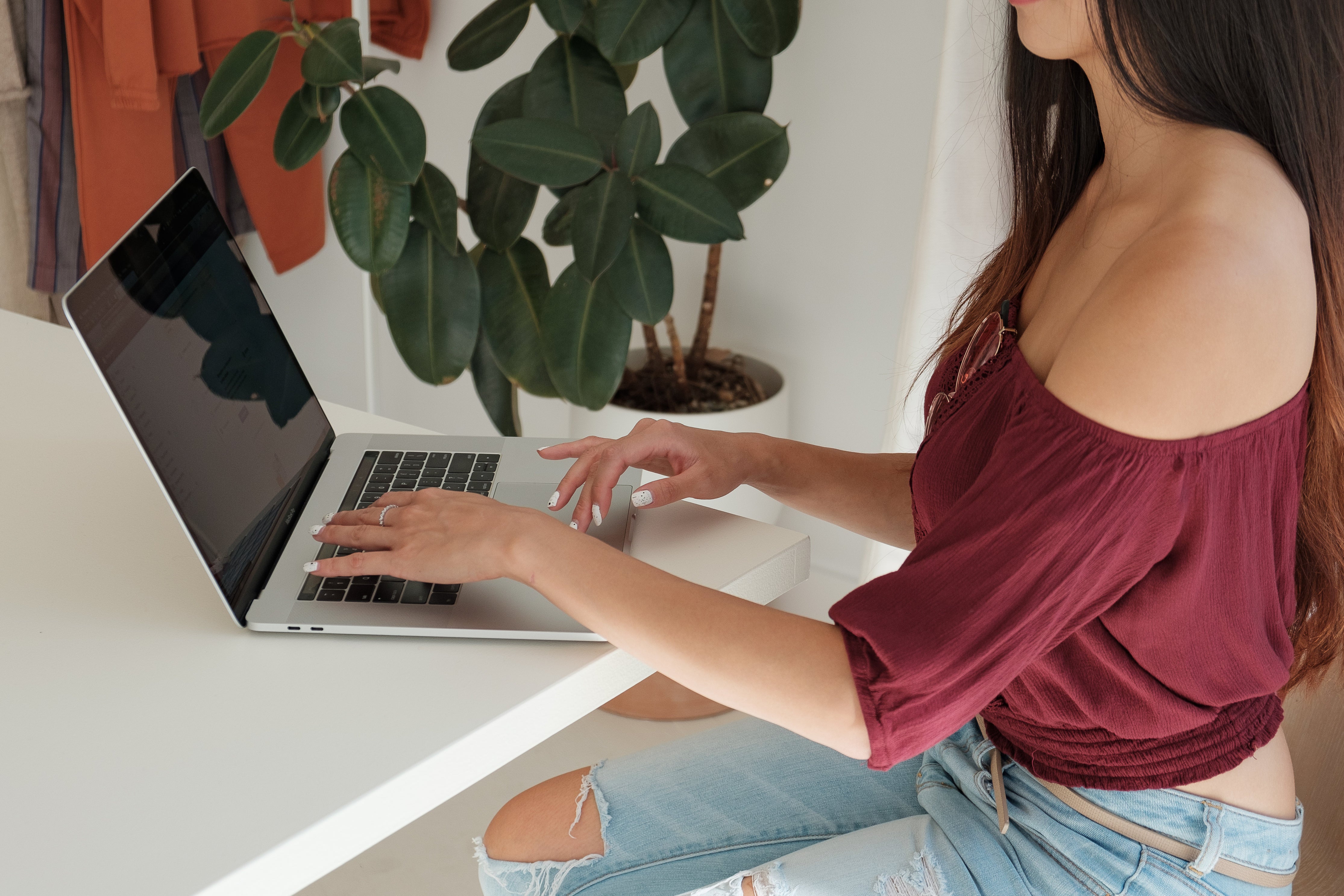 Blick von der Seite auf einen weißen Tisch an dem eine junge Frau in roten Oberteil vor einem aufgeklappten Laptop sitzt und etwas tippt. Die erste Funktion eines Online-Shop-Warenkorbs ist die Übersicht und mögliche Modifikation der Artikel. 