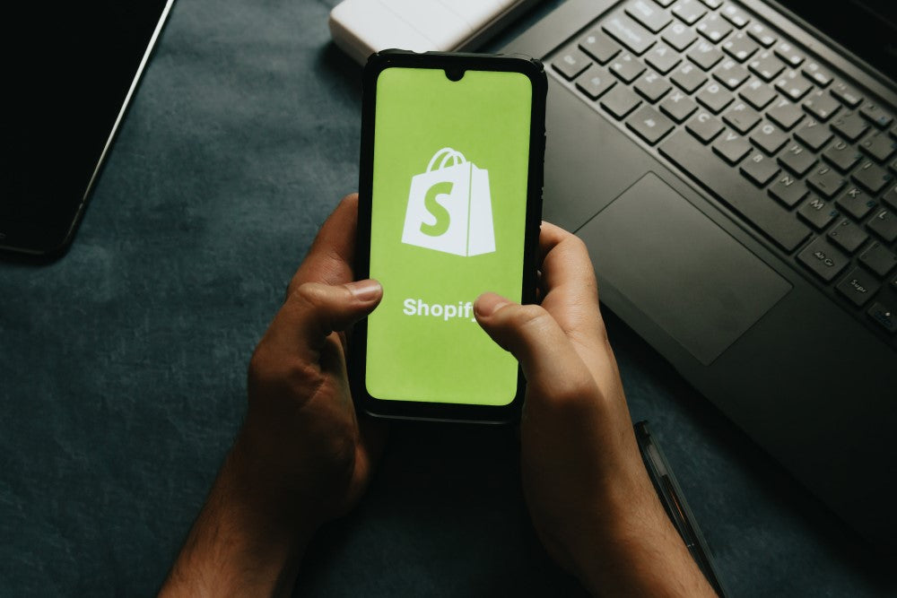 Gezeigt ist eine Person, die ein Handy in der Hand hält. Auf dem Bildschirm des Handys ist das Shopify Logo zu sehen.