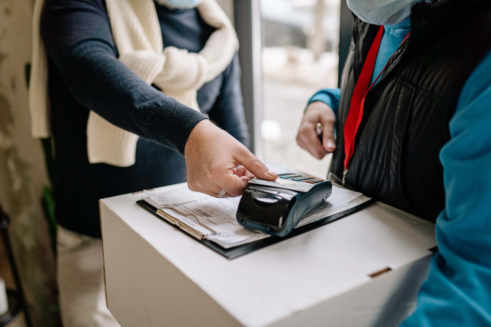 Zwei Personen deren Gesichter vom Bild abgeschnitten sind, stehen sich gegenüber. Die rechte Person hält ein großes Packet in der Hand auf dem ein Klemmbrett mit diversen Zetteln und ein Kartenlesegerät liegen. Die linke Person hält eine Karte an das Lesegerät. Erfahre in diesem Beitrag mehr über die Funktion von POS-Terminals!