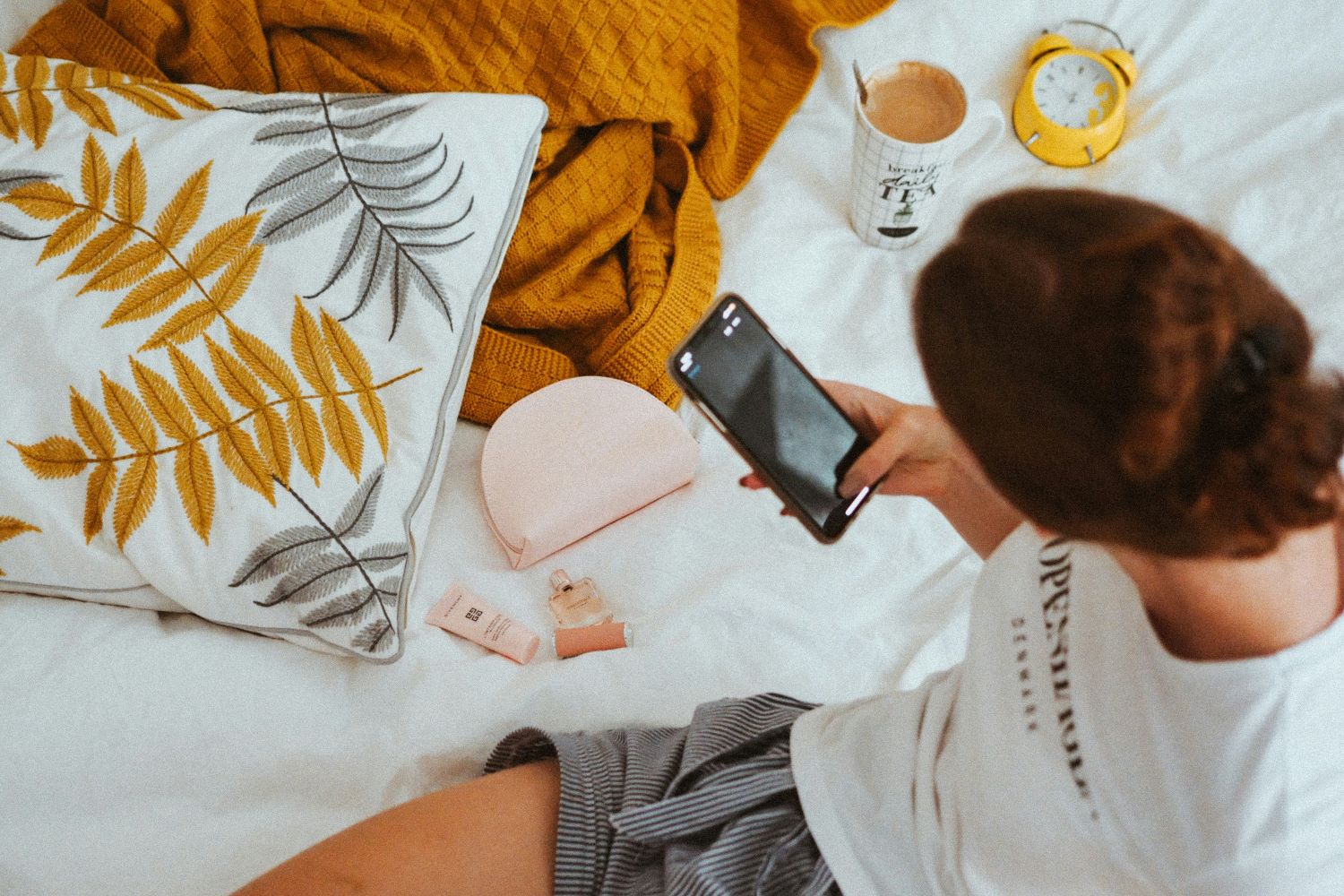 Gezeigt ist eine Frau, die auf einem Bett sitzt und mit ihrem Handy Bilder von Beauty-Produkten macht.