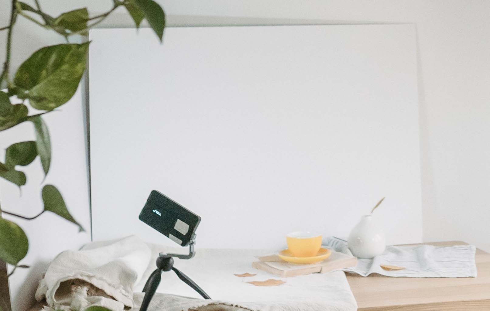 Auf einem Tisch steht eine weiße Platte an die dahinterliegende Wand gelehnt, während Smartphone und weitere Gegenstände zum Schmuck fotografieren bereitstehen