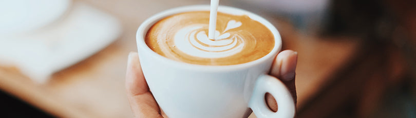Nahaufnahme einer Kaffeetasse in der Hand einer Person in die gerade Milch gegossen wird. Kaffee ist deine Leidenschaft? Mit diesem Hobby Geld verdienen ist vielleicht einfacher, als du denkst!