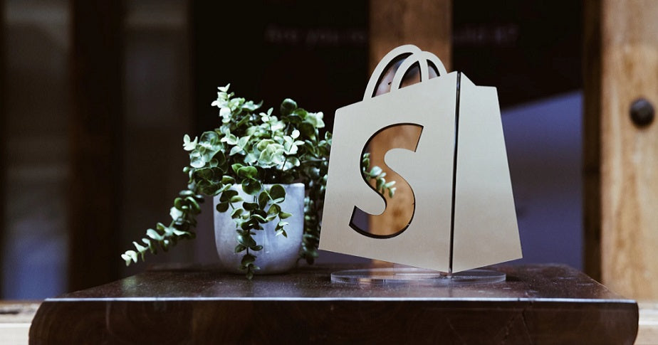 Die Figur eines Shopify-Logos steht zusammen mit einer Pflanze auf einem schwarzen Podest.