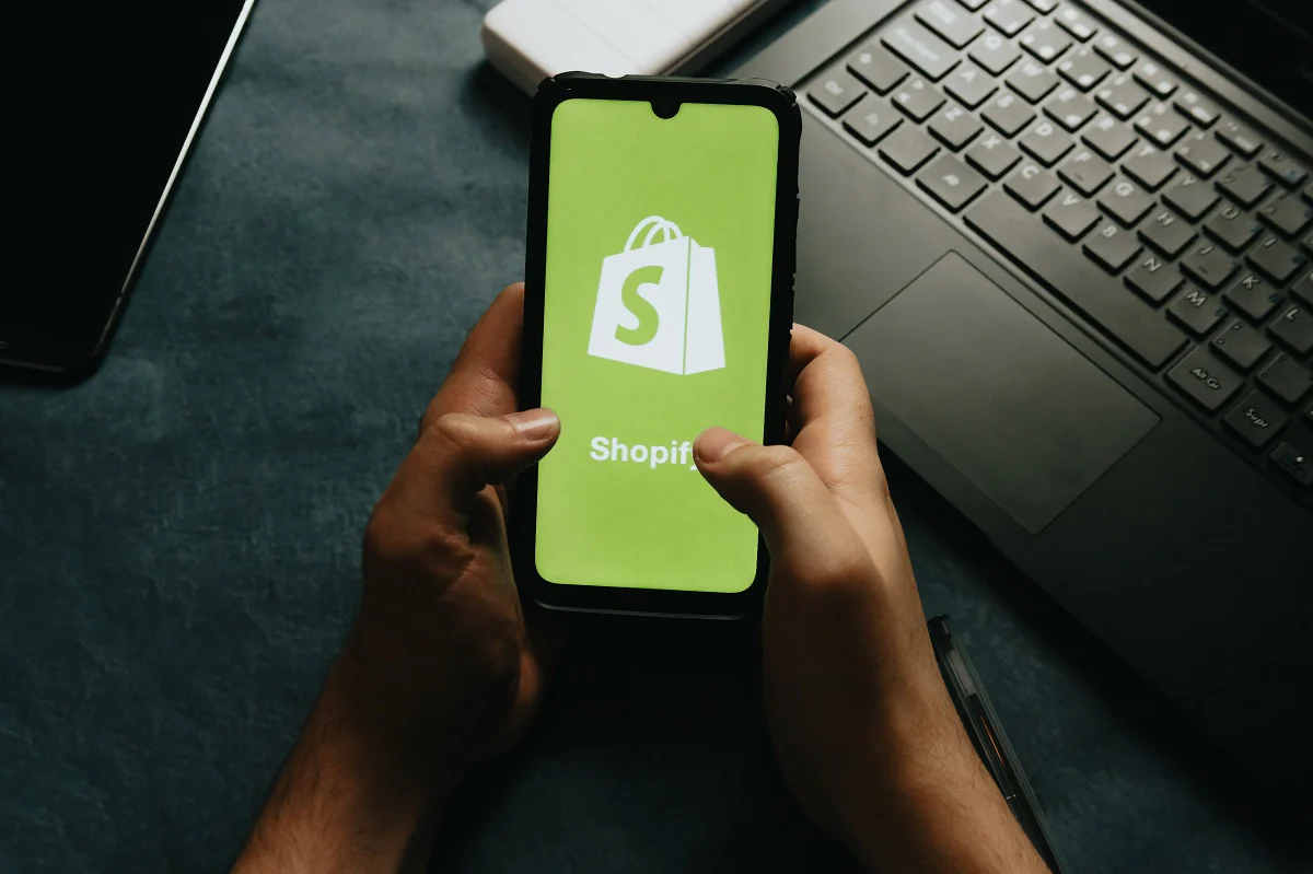 Das Foto zeigt zwei Hände, die ein Smartphone in der Hand halten auf dem die Startseite von Shopify aufleuchtet.