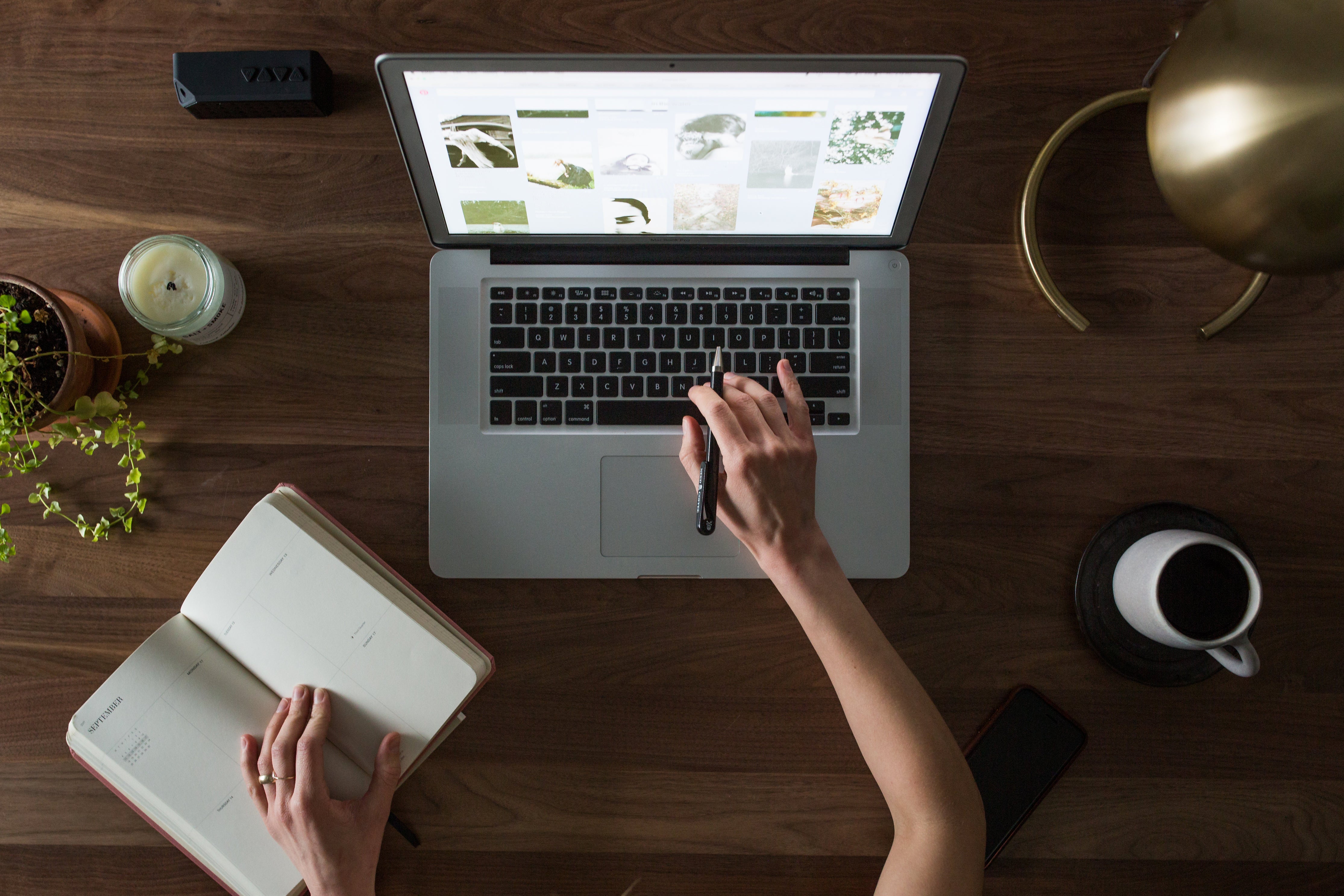 Blick von oben auf einen Schreibtisch auf dem ein aufgeklappter Laptop steht. Links daneben stehen eine Zimmerpflanze und eine Kerze. Rechts neben dem Laptop stehen eine Tasse Kaffee und eine Schreibtischlampe. Eine Person tippt mit einer Hand auf der Tastatur, die andere Hand liegt auf einem aufgeschlagenen Notizbuch. Die erfolgreiche Unternehmensgründung fußt auf einem durchdachten Businessplan. Wir zeigen dir, was du beachten solltest!