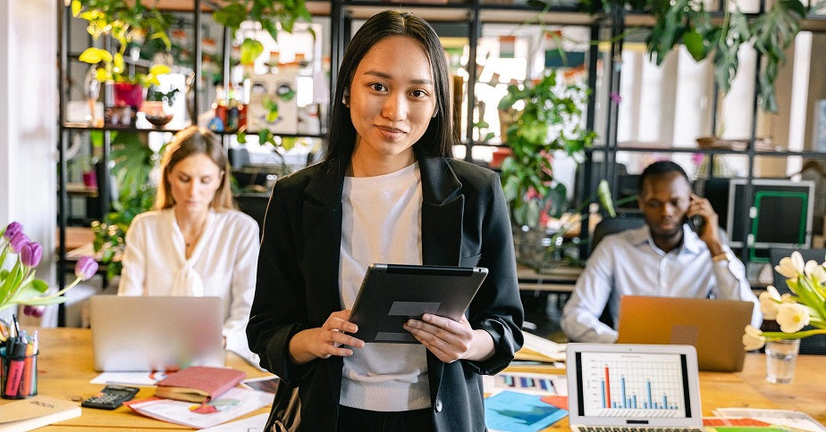 Eine junge Frau steht in einem hellen Büro mit einem Tablett in der Hand und lächelt in die Kamera. Hinter ihr sitzen zwei Personen am Schreibtisch und arbeiten. Dein eigenes Unternehmen gründen kann eine echte Herausforderung sein. Damit diese dir gelingt, haben wir dir einige Tipps aufgeschrieben!