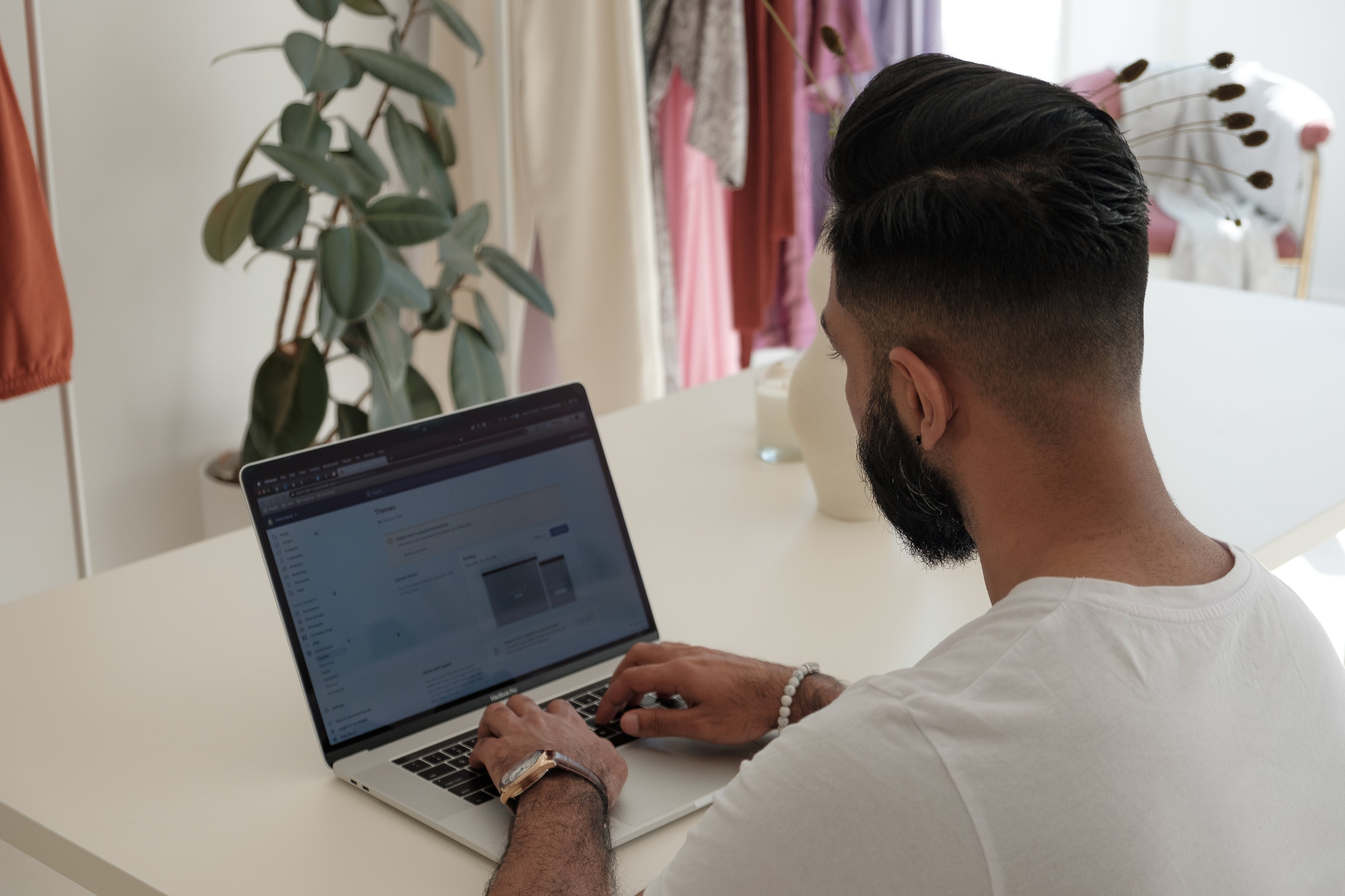 Blick über die Schulter eines jungen Mannes, der an einem weißen Schreibtisch sitzt und etwas auf einem aufgeklappten Laptop tippt. Im Hintergrund sind eine Zimmerpflanze und ein Kleiderständer zu sehen. Der Bildschirm des Laptops zeigt das Backend von Shopify. Die richtige Content-Marketing-Strategie hilft dir bei der Vermarktung deines Onlineshops.