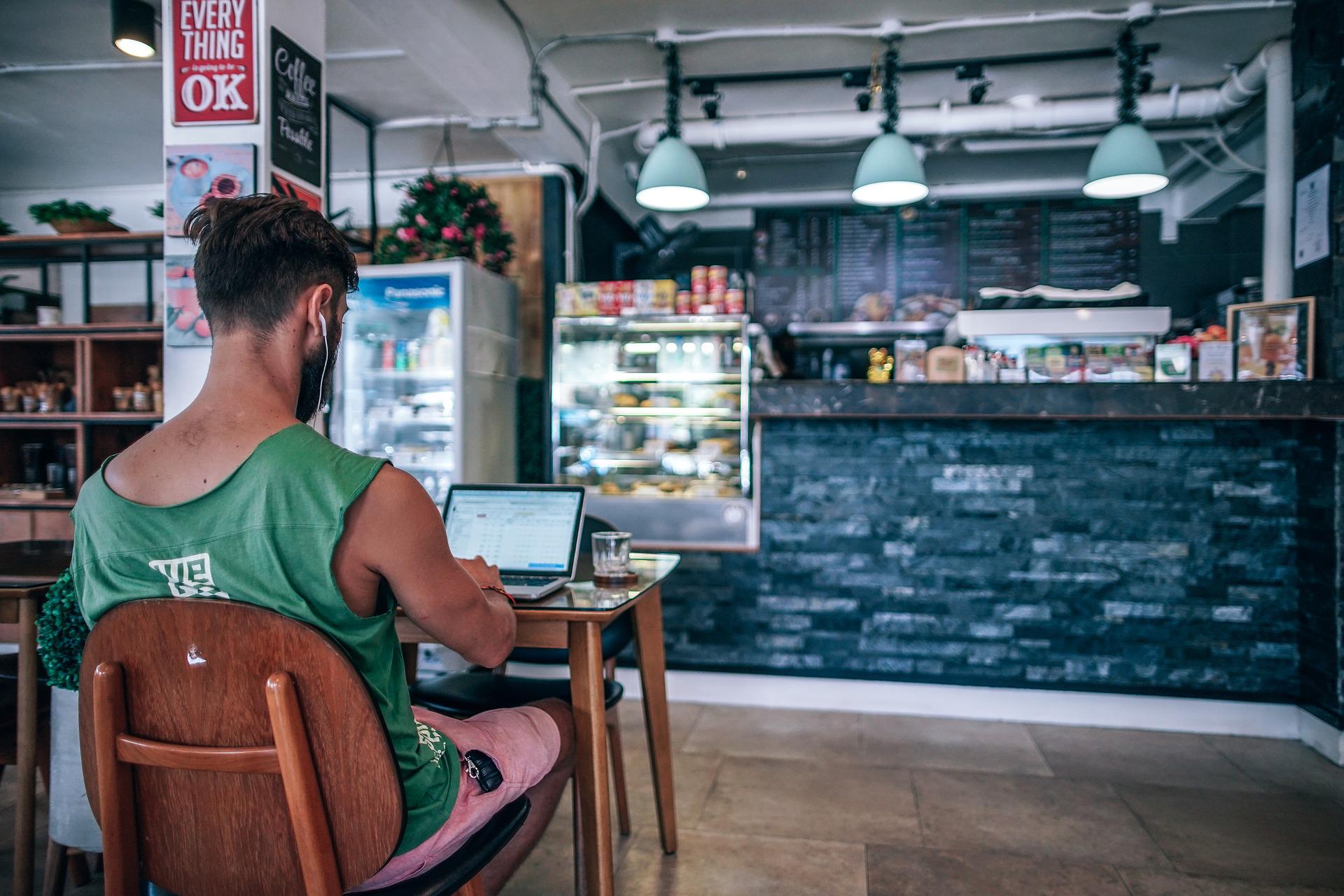 Blick auf eine Theke eines Cafés. Im Vordergrund sitzt ein junger Mann an einem braunen Holztisch mit einem aufgeklappten Laptop vor sich. Für den ersten Vertrauensvorschub solltest du dich auf Empfehlungen und Kooperationen konzentrieren, um deinen Onlineshop erfolgreich zu vermarkten.