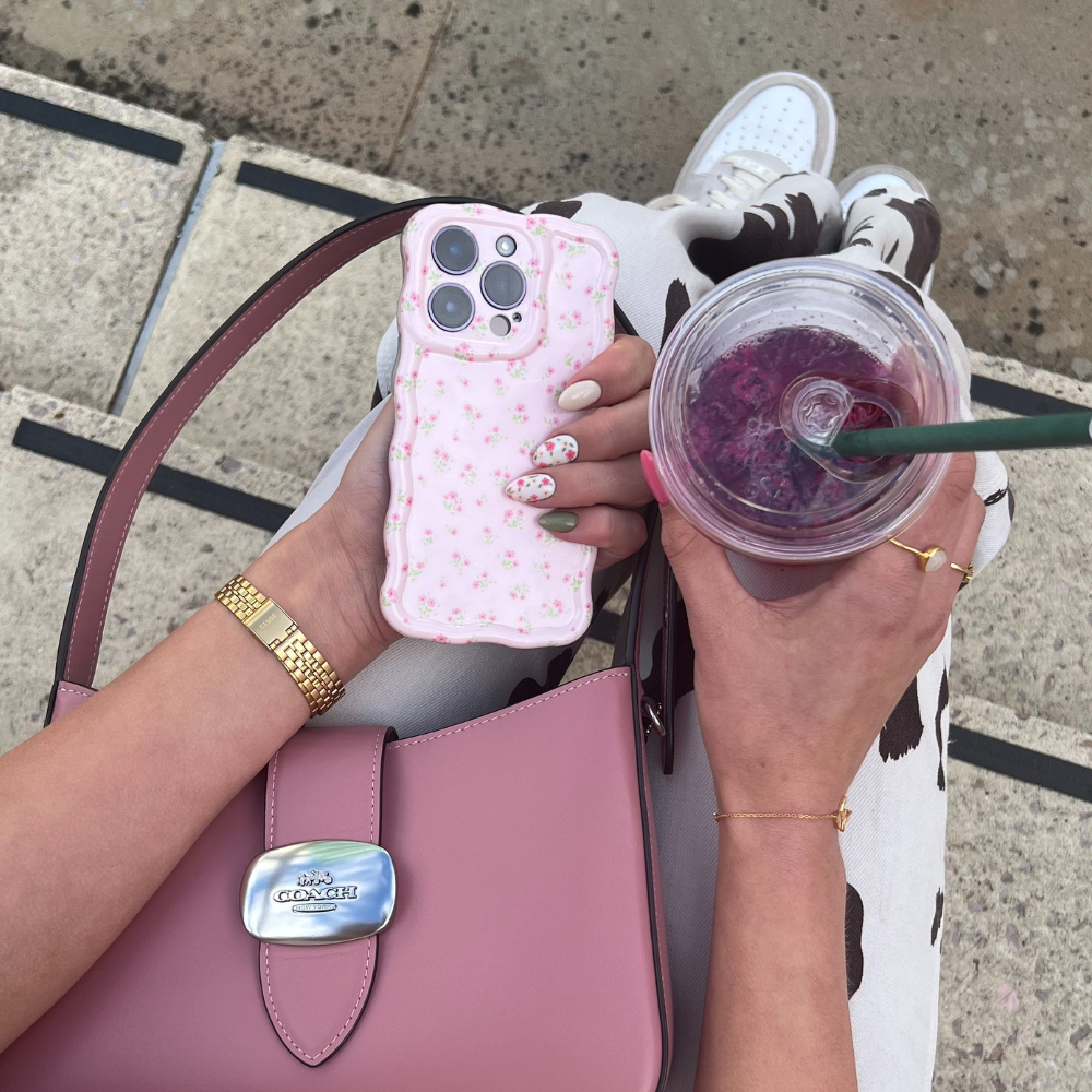 Hands holding a floral phone case and a pink drink, with a pink handbag on lap.