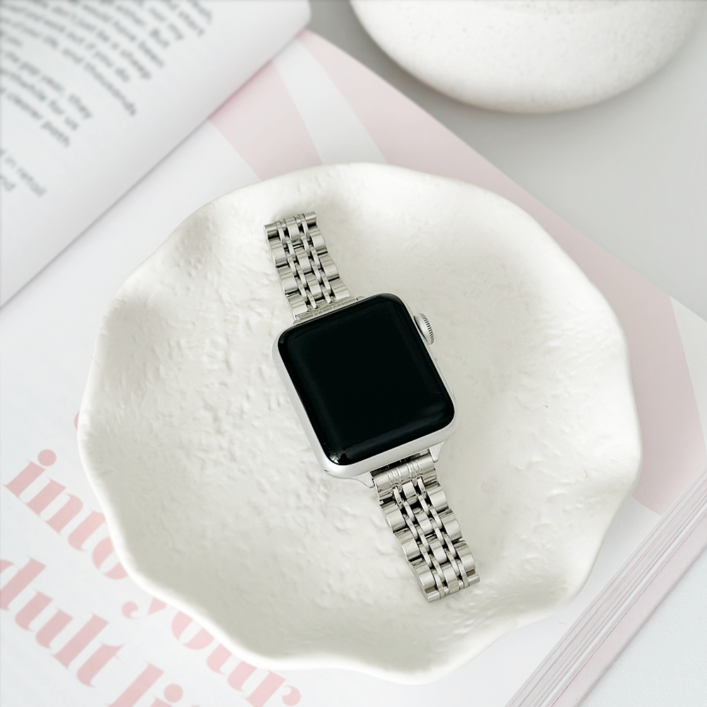 Smartwatch with a metal band resting in a white dish next to an open book.