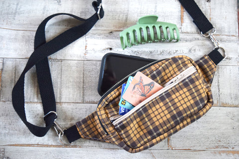 A brown plaid crossbody bag with cards, and iPhone and a hair clip on a gray wooden background.