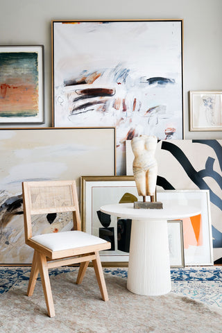 On the back wall are various abstract art pieces on canvas. Most of the paintings are neutral color except the one on the top left (it has a bright orange and deep blue). In the foreground is a wicker chair and white table. On top of the table is a white sculpture of a woman's torso.