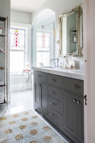 A naturally lit bathroom with dark cabinetry and a stained glass window in the corner. The sink has silver fixtures and a classic marble countertop with grey striations. There is a small blue grey rug to stand on while washing your hands. The rug has flecks of yellow stripes and yellow flower-like geometric designs.