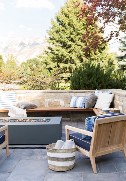 Wooden patio furniture surrounds a fire table. Large trees are in the background.