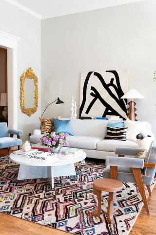 The focal point of the room is a large warm toned multicolored geometric shaped rug. On top of the rug is a low white circular coffee table and a large white couch. On the back walls is an ornate gold mirror and a portrait of a woman in a black outline.
