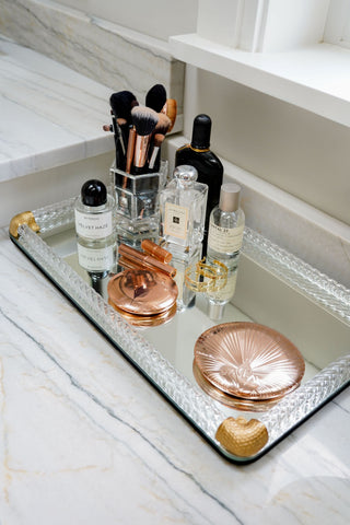 A decorative mirror tray has gold on the corners. The tray is holding makeup brushes, cosmetics, and a compact.