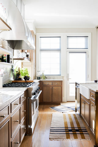 A brightly lit kitchen with large windows on the back wall and an aisle with cabinets on either side. On the right is a sink and a small plaid area rug that is mostly brown.